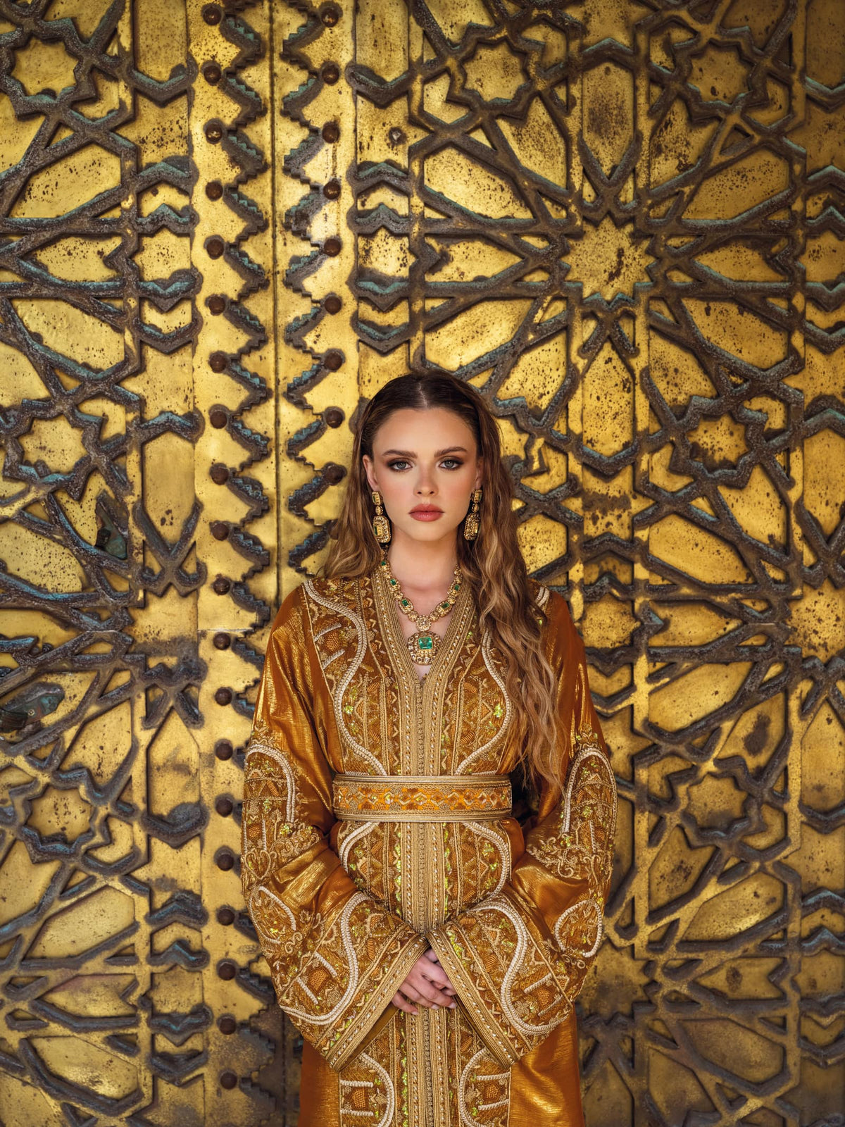 A bronze Moroccan caftan with gold embroidery, photographed before a carved brass door.